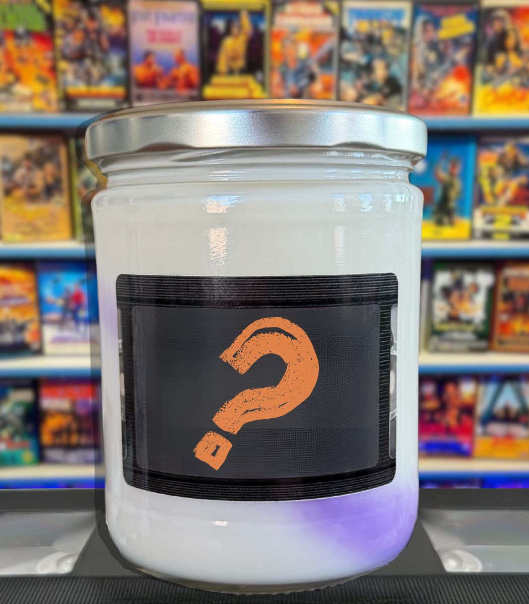 A large white candle in a clear glass jar with a silver metal lid, featuring a black label with an orange question mark. The background shows blurred shelves filled with colorful books or boxes.